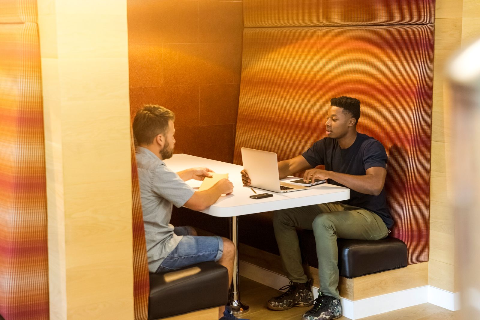 Dois homens conversam com notebook e anotações em uma cabine de reunião acústica.