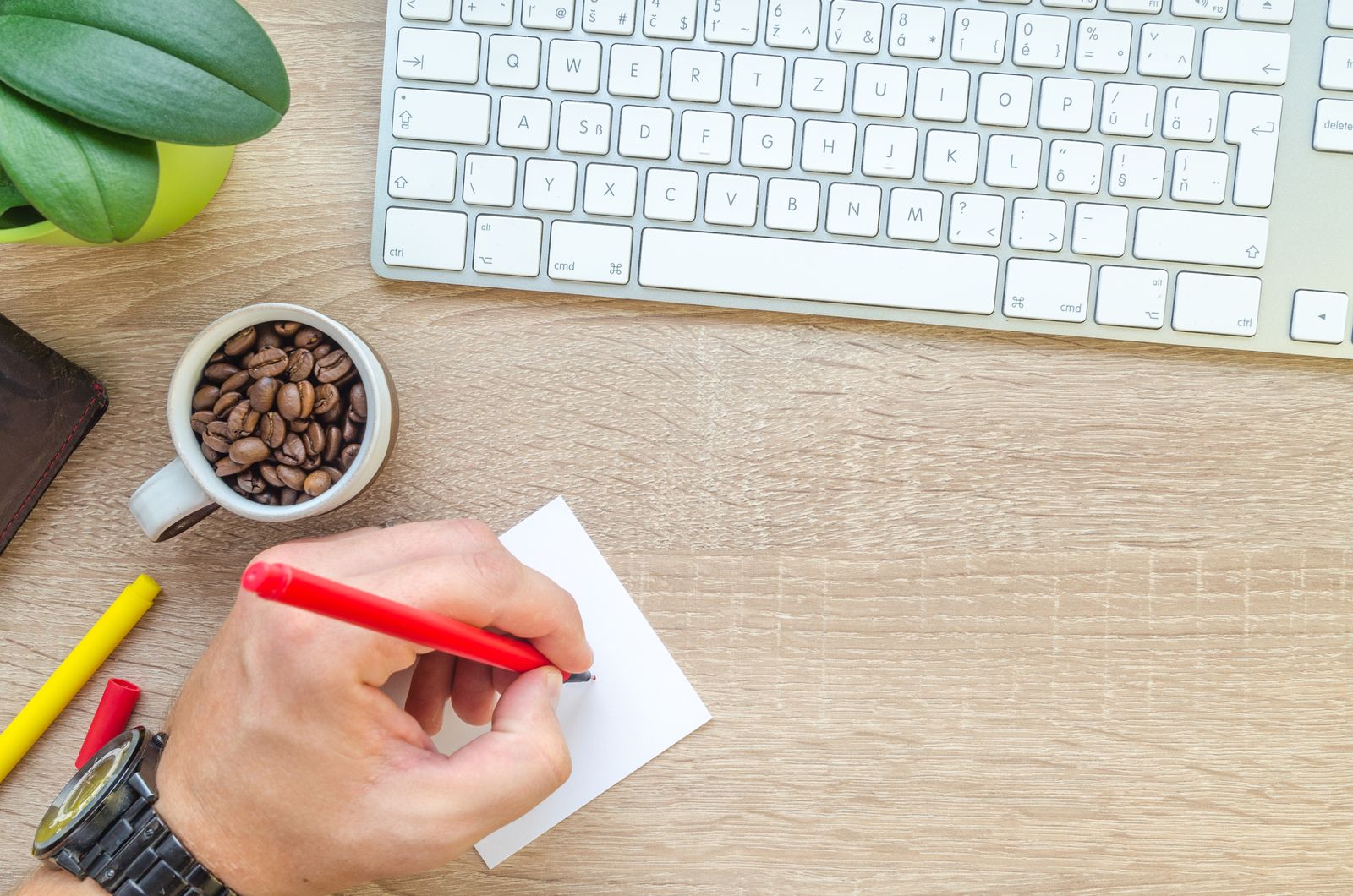 Vista superior de uma mesa de trabalho com teclado, caneta, xícara de grãos de café e mão escrevendo em um papel.