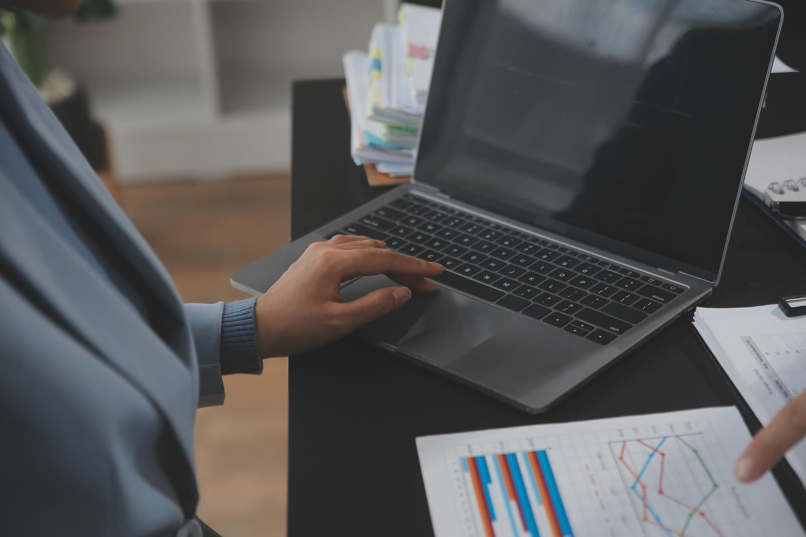 Pessoa digita em um notebook sobre mesa preta ao lado de relatórios com gráficos impressos.