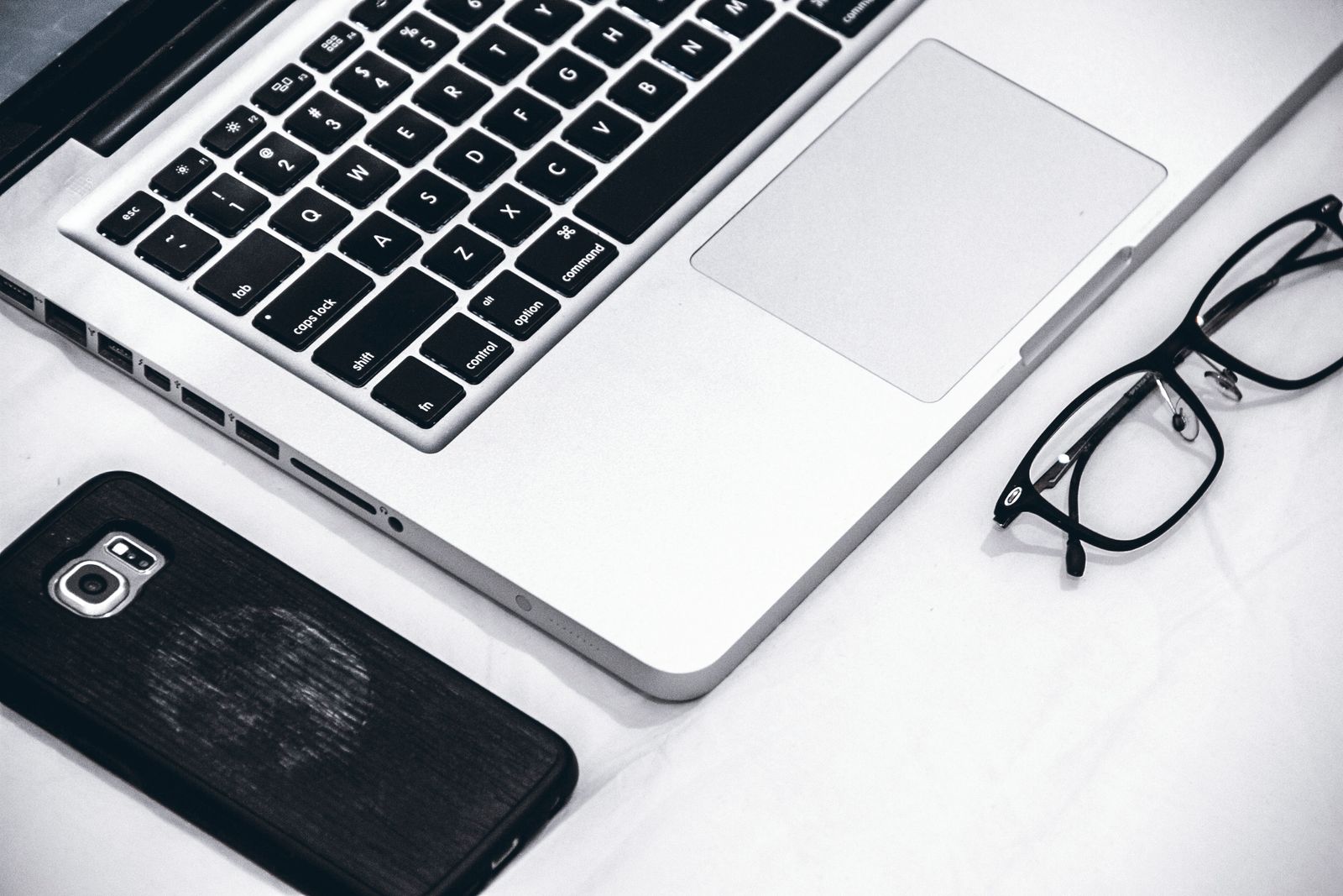 Notebook, celular e óculos sobre uma superfície branca, em preto e branco.